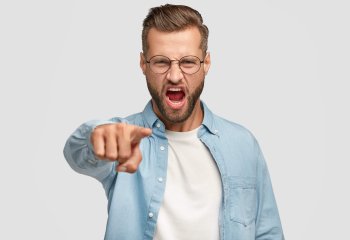 Outraged bearded guy points at camera, blames somebody in wrong doing, has desperate expression, dressed in blue shirt and spectacles, isolated over white background. Negative emotions concept