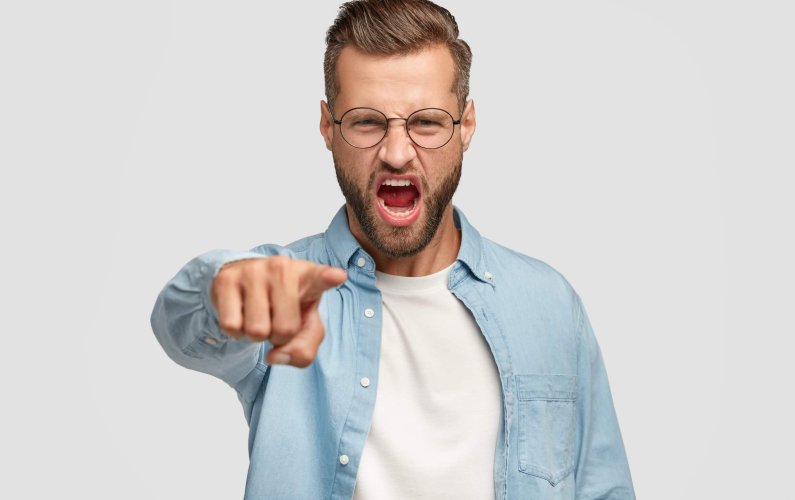 Outraged bearded guy points at camera, blames somebody in wrong doing, has desperate expression, dressed in blue shirt and spectacles, isolated over white background. Negative emotions concept